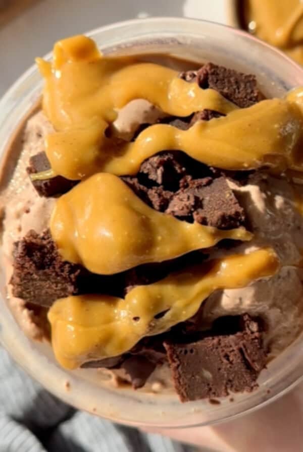 A bowl of chocolate ice cream topped with caramel sauce and chunks of chocolate brownies. A gold spoon is partially visible on the right.