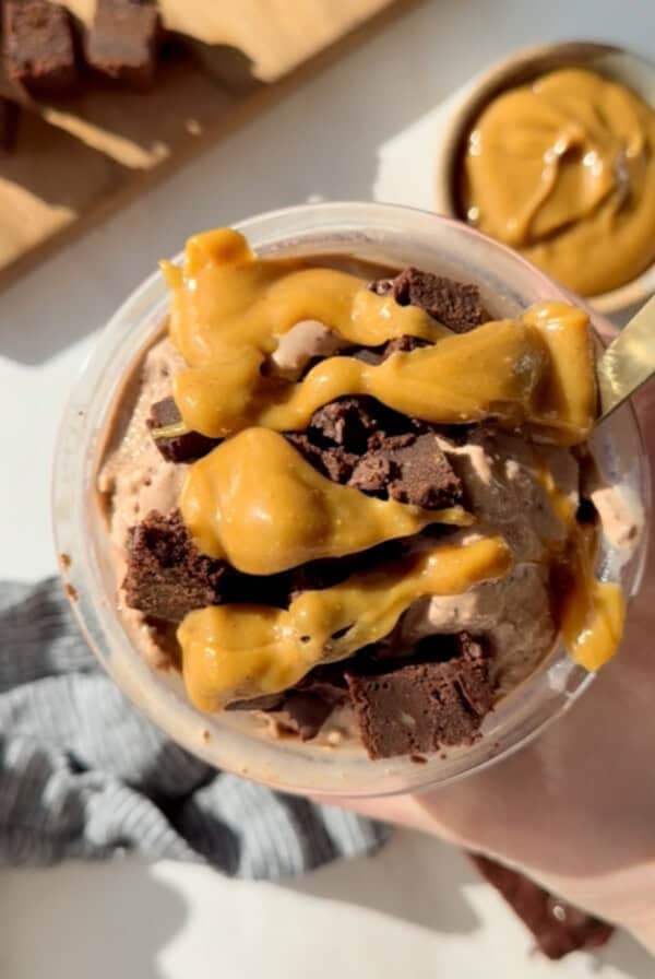 A cup of chocolate ice cream topped with caramel sauce and brownie chunks, with a spoon in it. A bowl with caramel is visible in the background.