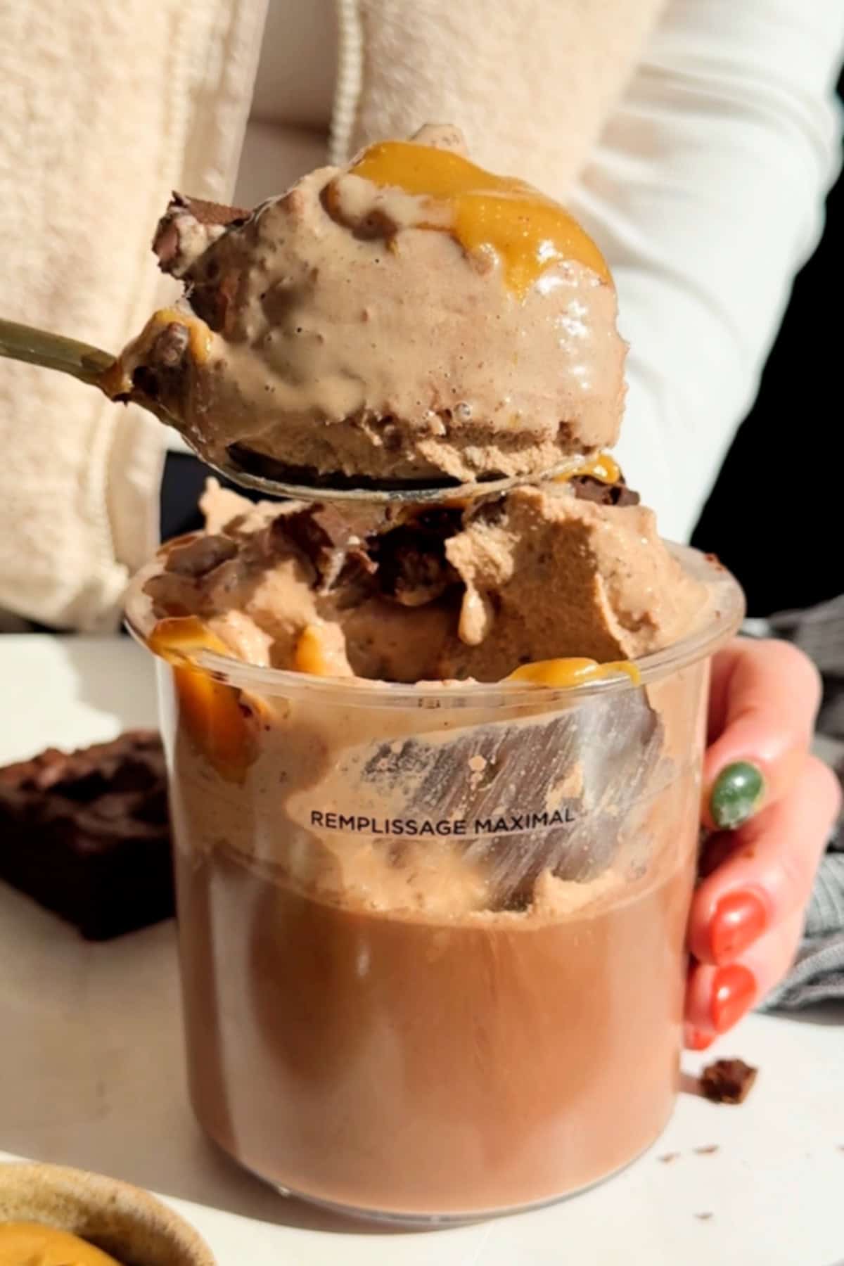 A person holds a spoon with ice cream drizzled with caramel over a clear cup, which seems filled to the maximum line. A brownie is visible in the background.