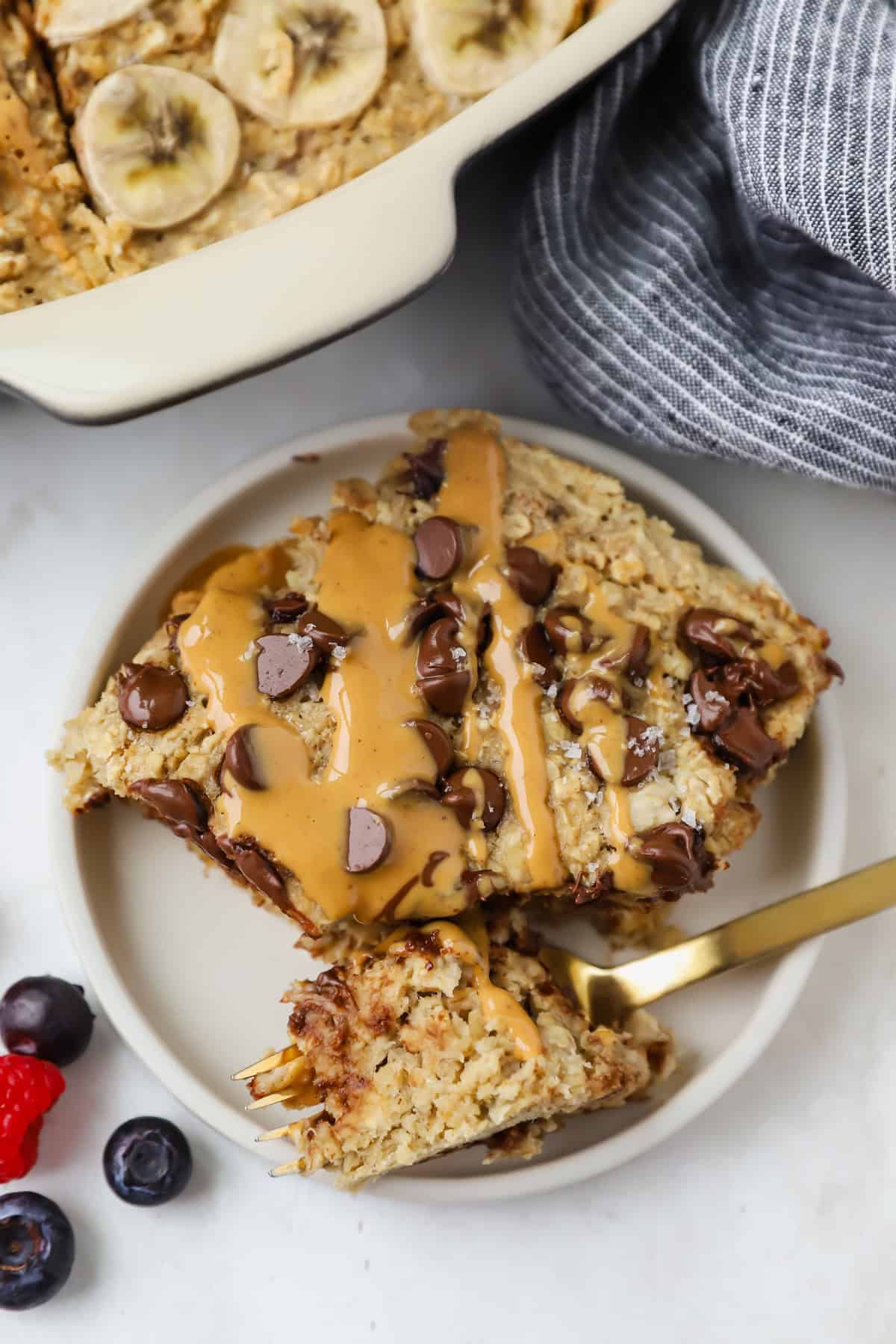 A plate with a slice of banana baked oatmeal, freshly baked, topped with chocolate chips and a peanut butter drizzle. A fork rests on the plate, accompanied by vibrant blueberries and a raspberry on the side.