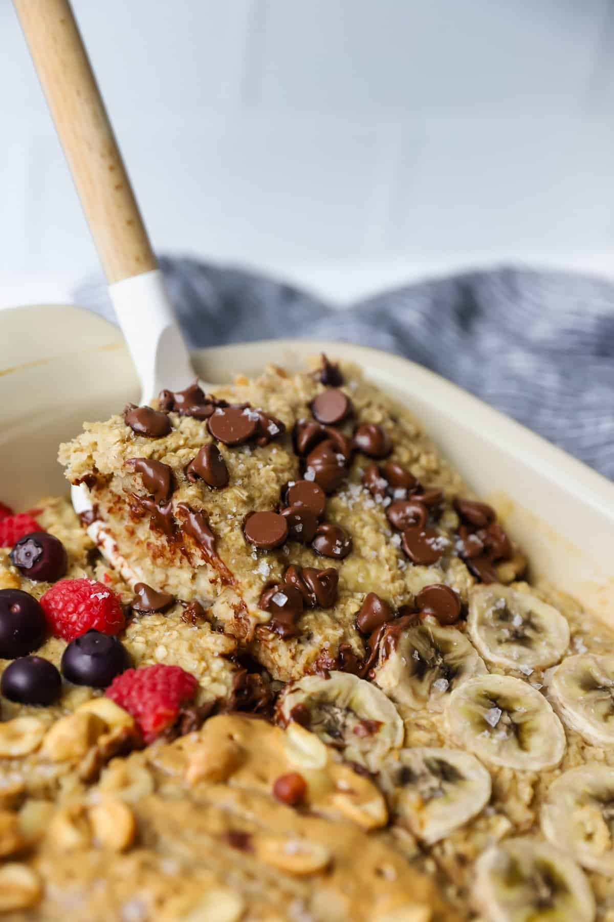 A delicious baked oatmeal, bursting with chocolate chips, sliced bananas, berries, and nuts. A spatula gracefully lifts a portion of this comforting dish.