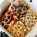 A dish of baked oatmeal is adorned with four tempting toppings: chocolate chips, banana slices, berries, and peanuts drizzled with peanut butter. A serving is being carefully lifted with a spatula, showcasing its delicious layers.