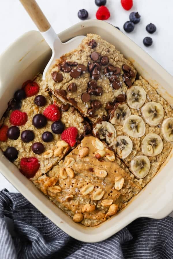 A baking dish with four sections of baked oatmeal topped with chocolate chips, banana slices, mixed berries, and peanut butter with peanuts. A spatula rests on one section.