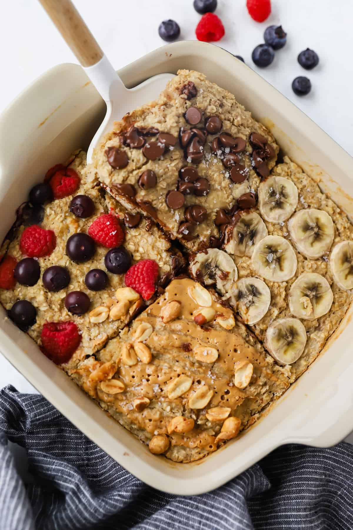 A baking dish with four sections of baked oatmeal topped with chocolate chips, banana slices, mixed berries, and peanut butter with peanuts. A spatula rests on one section.