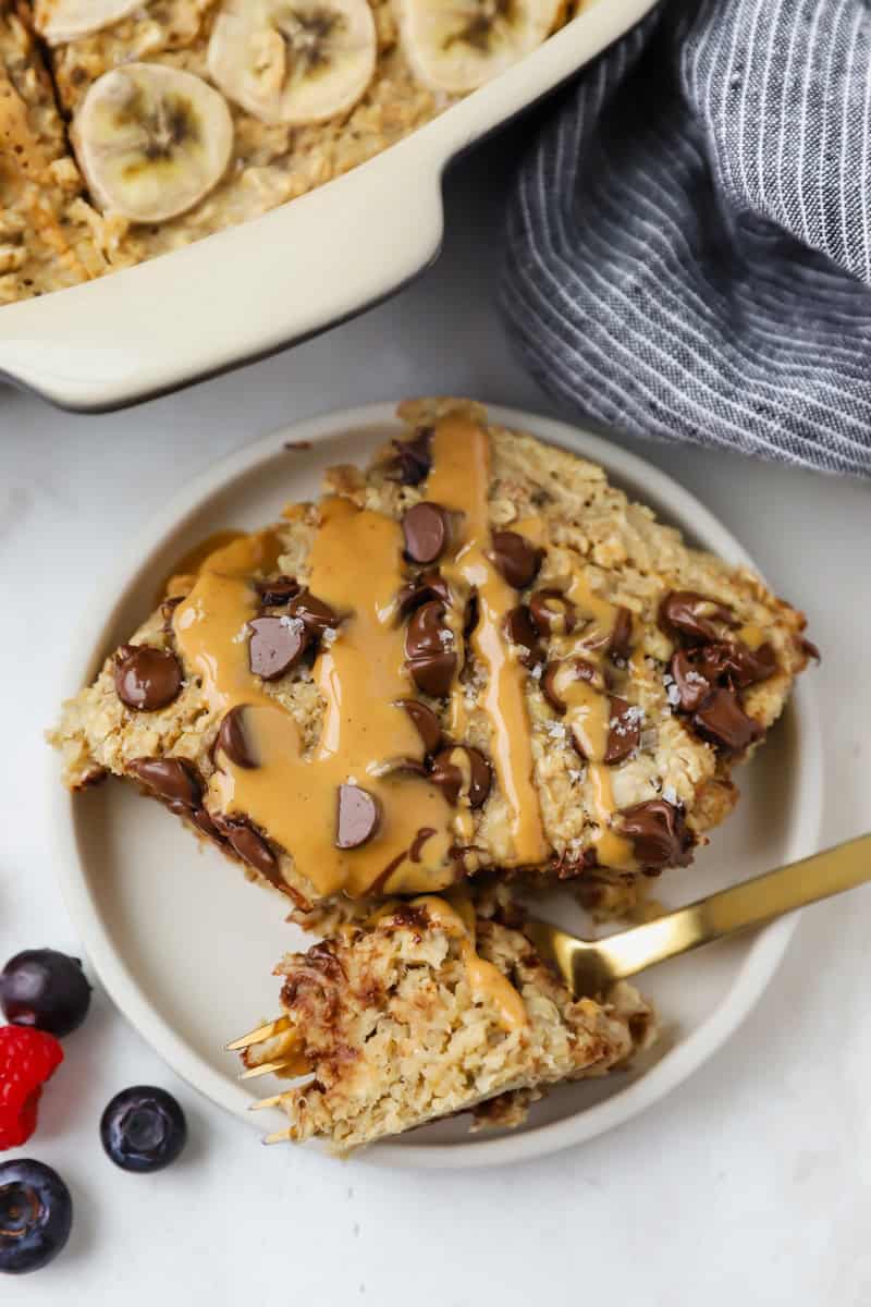 A plate with a slice of baked oatmeal topped with chocolate chips and peanut butter sits next to a casserole dish, fresh berries beside the plate, and a striped cloth in view.