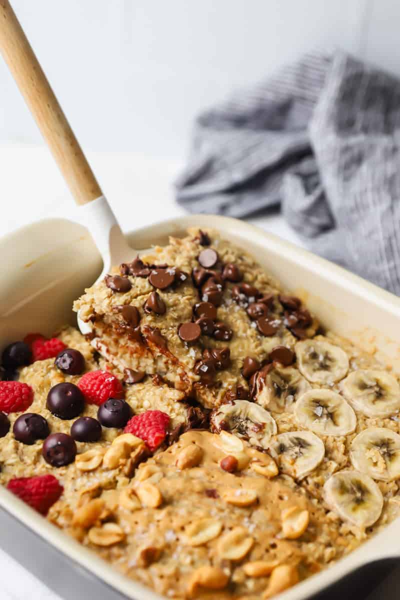 In a dish, the baked oatmeal is divided into sections adorned with chocolate chips, banana slices, berries, and nuts. A serving has been scooped out with a spatula.