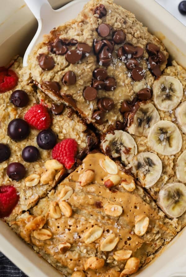 Baked oatmeal in a dish with four sections, each crowned with delicious toppings: chocolate chips, banana slices, mixed berries, and peanuts. A spatula lifts one tempting portion of the baked oatmeal.