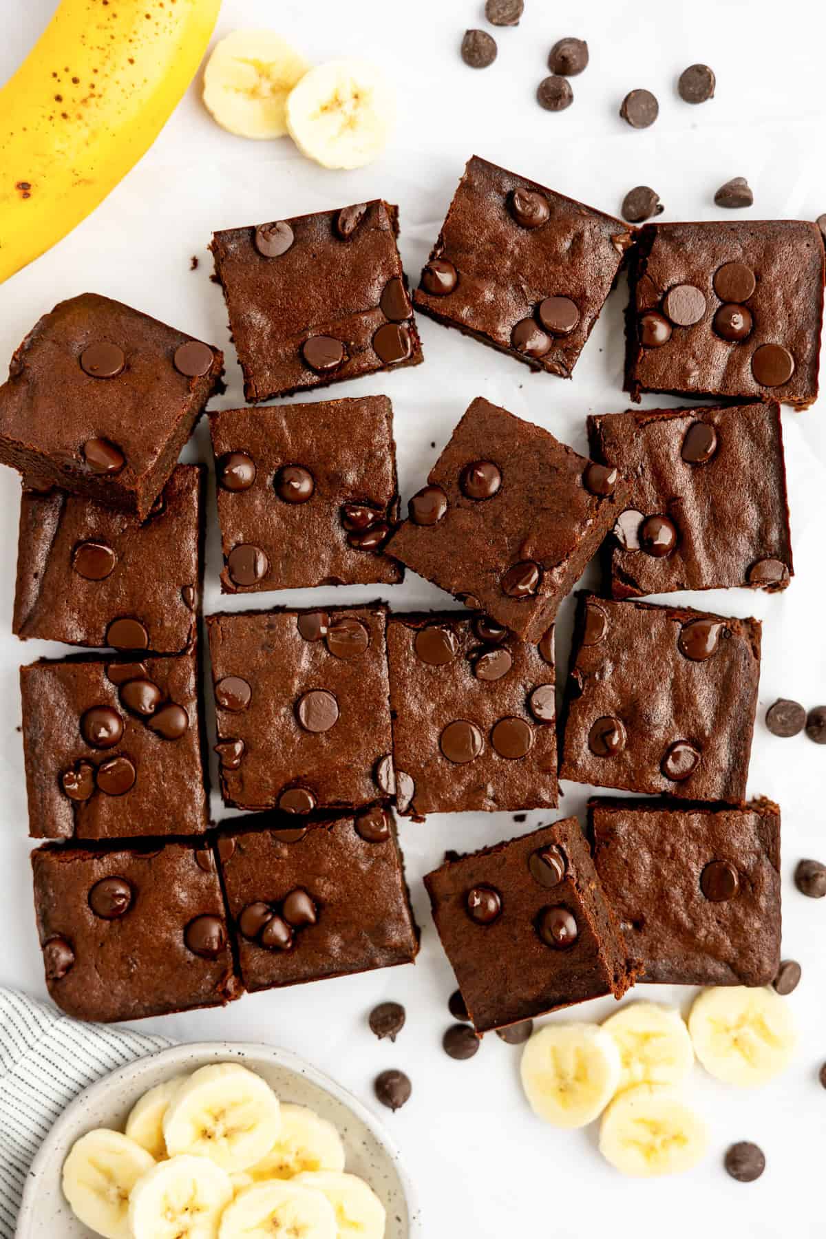 Brownies with chocolate chips arranged on a surface, surrounded by banana slices and a whole banana.