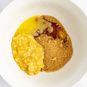 A white bowl containing mashed bananas, brown sugar, and melted butter on a white surface.