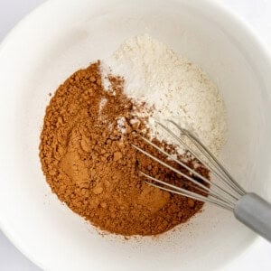 A white bowl with cocoa powder, flour, and baking soda being mixed with a metal whisk.