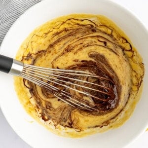 A large white bowl with a whisk, mixing ingredients that include a swirled liquid and batter, next to a whole banana on a white surface.