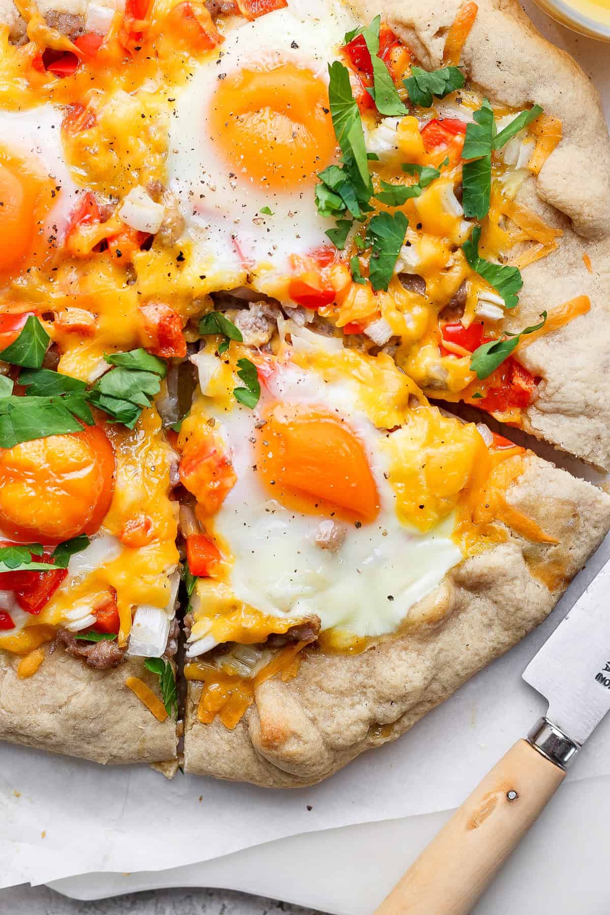 A pizza topped with sunny-side-up eggs, melted cheese, diced red bell peppers, onions, and fresh herbs. A slice is cut out, with a knife resting nearby.