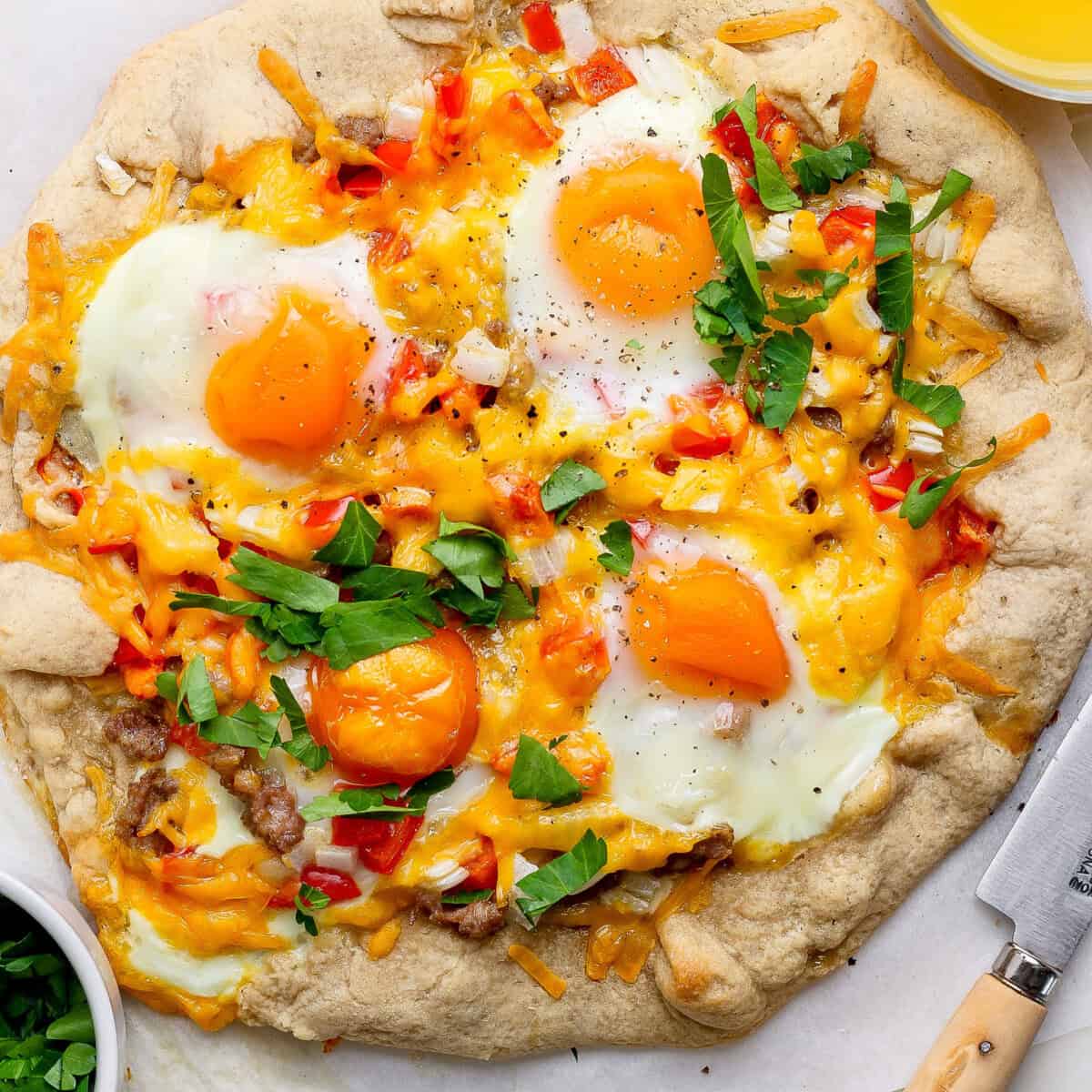 A rustic breakfast pizza topped with sunny-side-up eggs, cheese, diced red peppers, and fresh parsley on a light crust, placed next to a knife and a bowl of chopped greens.