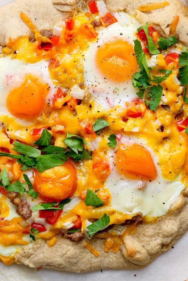 A rustic breakfast pizza topped with sunny-side-up eggs, cheese, diced red peppers, and fresh parsley on a light crust, placed next to a knife and a bowl of chopped greens.