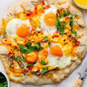 A baked breakfast pizza topped with sunny-side-up eggs, cheese, red peppers, and herbs on parchment paper, with a bowl of chopped parsley and a glass of orange juice nearby.