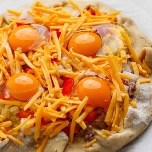 An unbaked pizza topped with raw eggs, shredded cheese, and chopped red peppers on a parchment-lined surface.