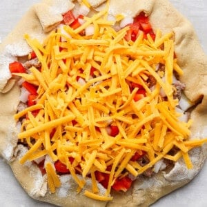 Unbaked pizza with cheddar cheese, diced red peppers, onions, and mushrooms on dough.