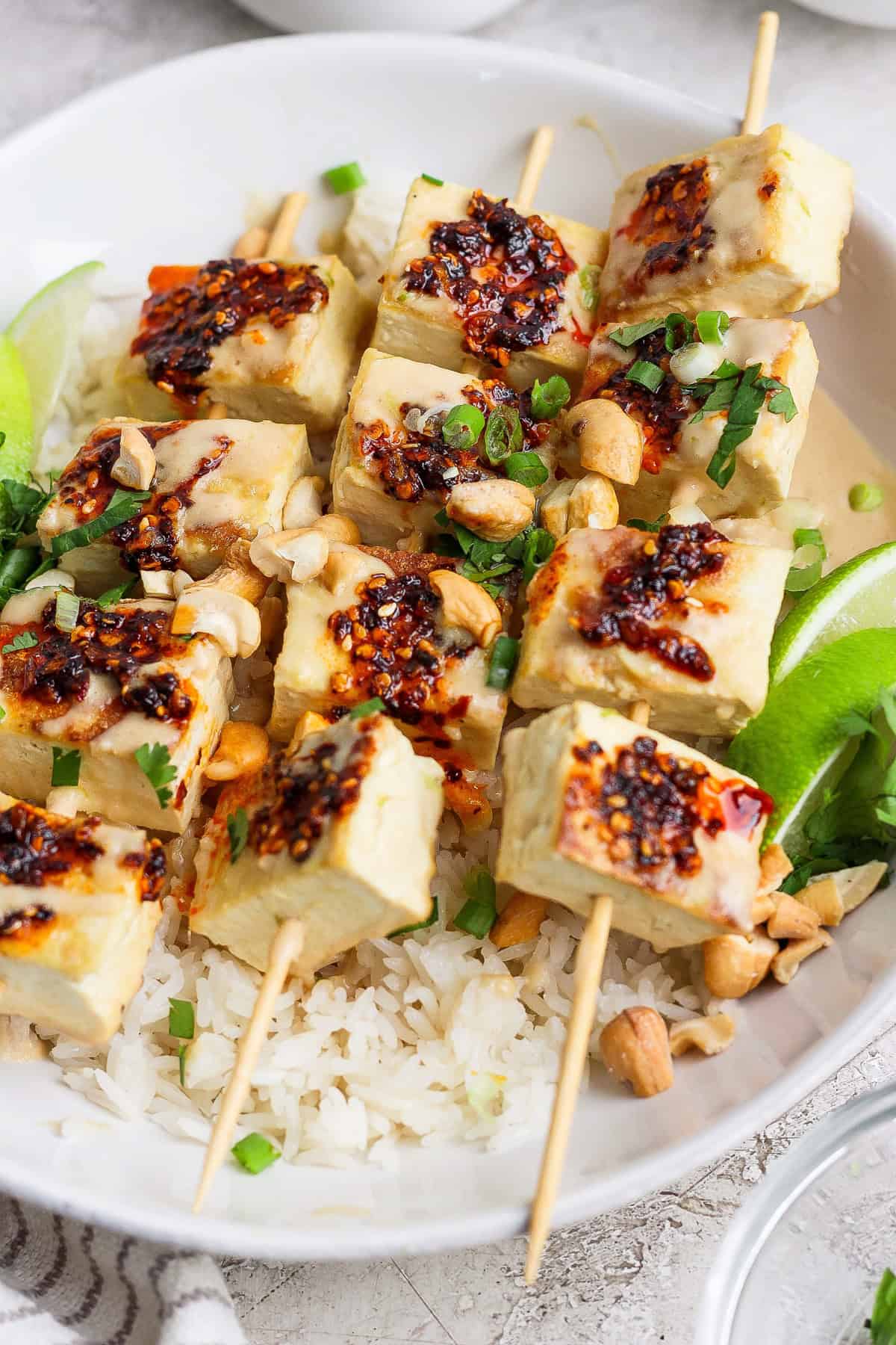 Skewered tofu cubes with chili sauce are placed over a bed of rice, garnished with chopped green onions, peanuts, and lime wedges.