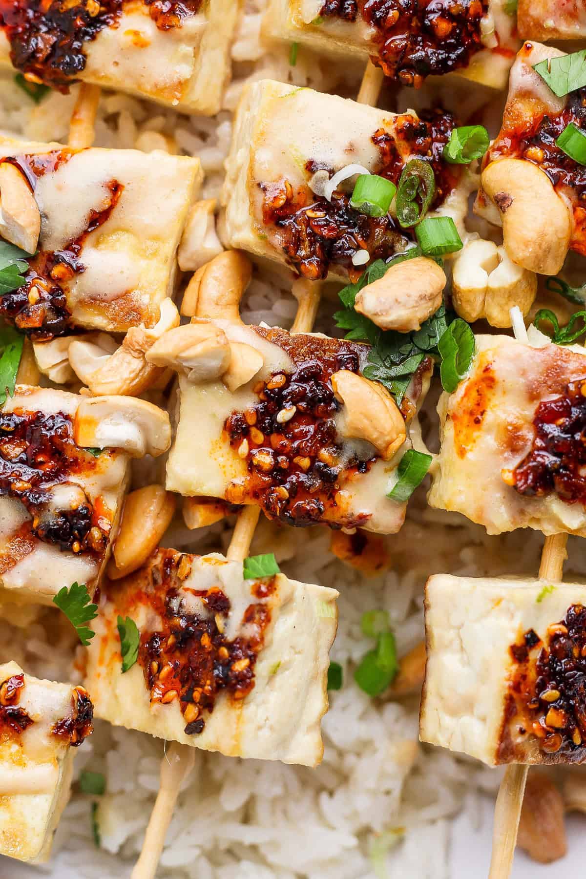 Grilled tofu skewers topped with chili sauce, garnished with green onions and cashews, served on a bed of rice.