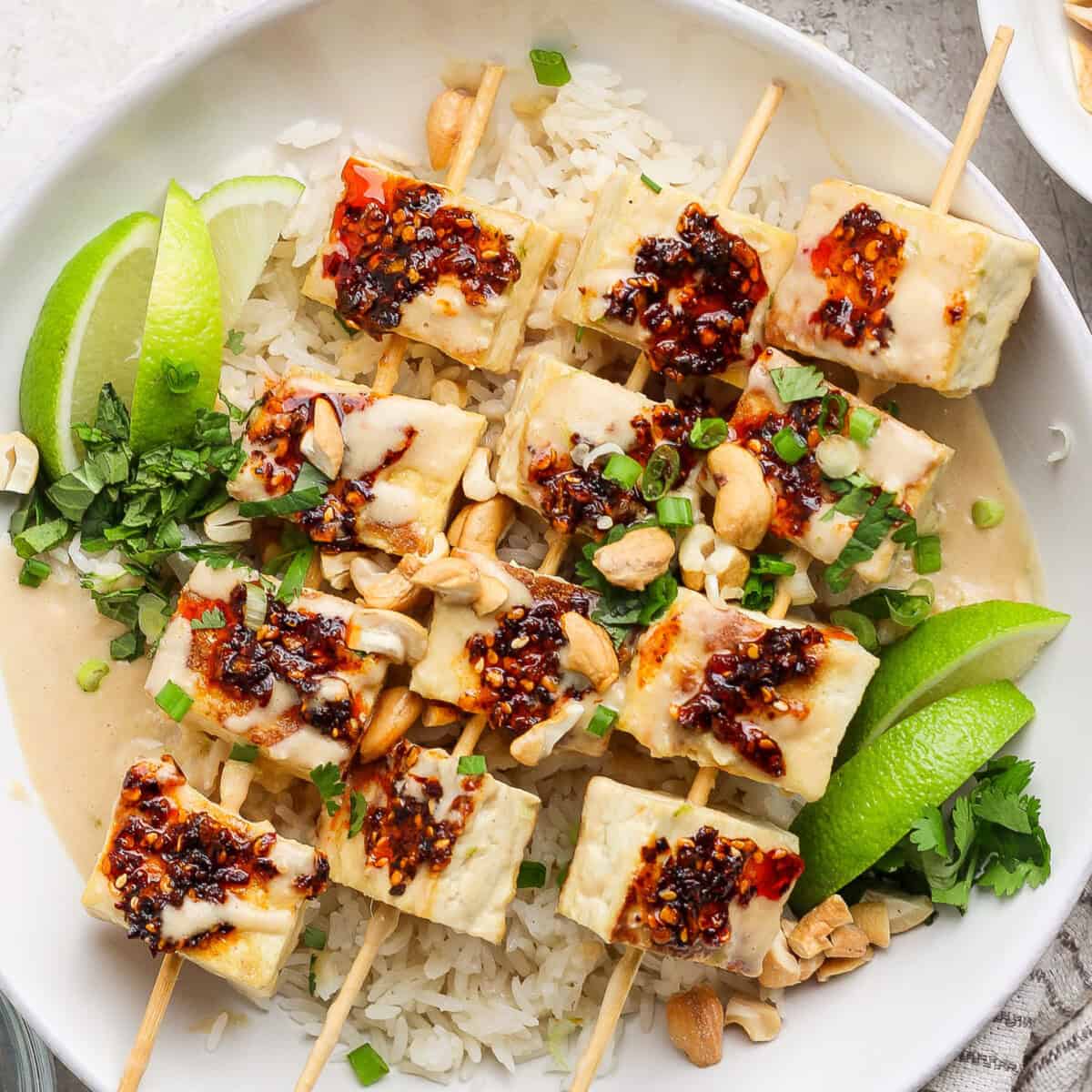 A dish with skewered tofu topped with chili sauce on rice, garnished with lime wedges, green onions, peanuts, and herbs.