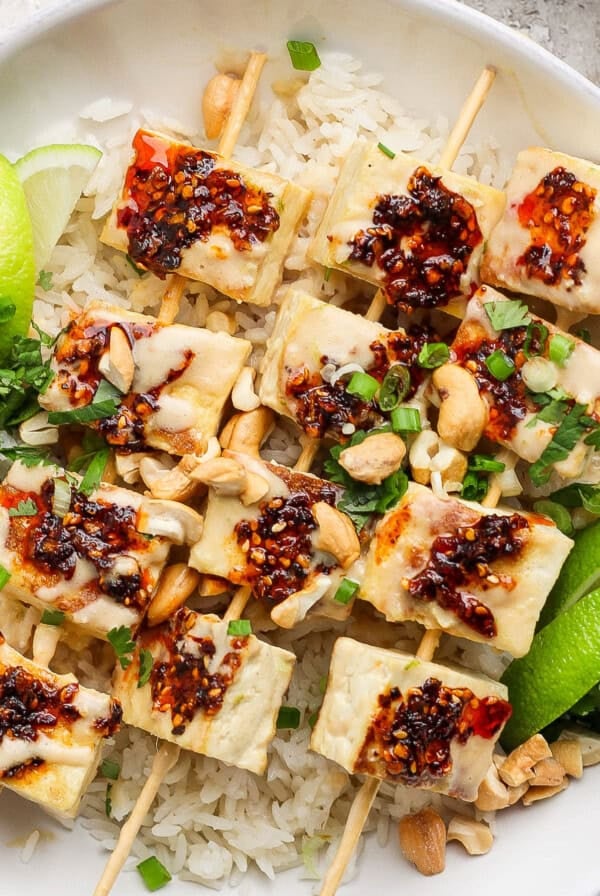 A dish with skewered tofu topped with chili sauce on rice, garnished with lime wedges, green onions, peanuts, and herbs.