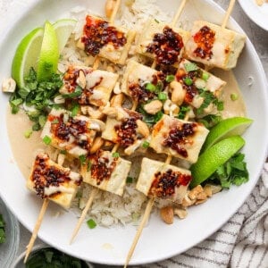 Tofu skewers with rice, garnished with lime wedges, cilantro, chopped nuts, and a drizzle of chili sauce, served in a white bowl.