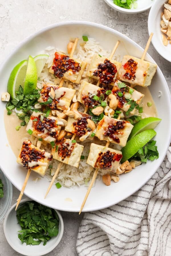 Tofu skewers with rice, garnished with lime wedges, cilantro, chopped nuts, and a drizzle of chili sauce, served in a white bowl.