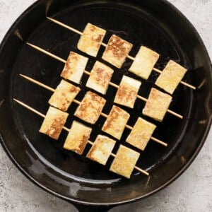 Skewered tofu pieces are grilled in a black cast iron pan on a textured light gray surface.