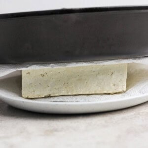 A block of tofu being pressed between a skillet and white plate, with paper towels underneath.