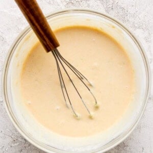 A glass bowl filled with a light beige batter. A whisk with a wooden handle rests in the batter. The surface beneath the bowl is textured and light-colored.