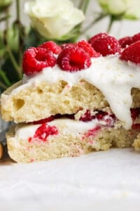 Close-up of a cake layered with white frosting and fresh raspberries, with a few white roses in the blurred background.