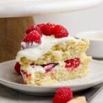 A slice of vanilla cake with cream and raspberries on a plate. A whole cake is partially visible in the background.