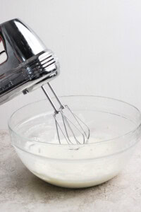 Electric hand mixer with beaters above a glass bowl of white liquid, possibly cream or batter, on a countertop.