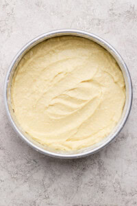 Cake batter spread evenly in a round, silver baking pan on a textured, light-colored surface.