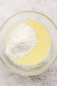 A glass bowl containing a mixture of yellow batter with a mound of white flour on top, set on a textured surface.