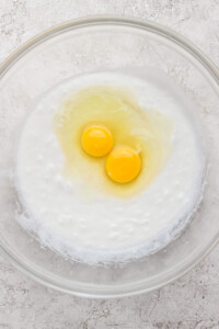 Two raw eggs cracked on top of a mixture in a clear glass bowl on a textured surface.