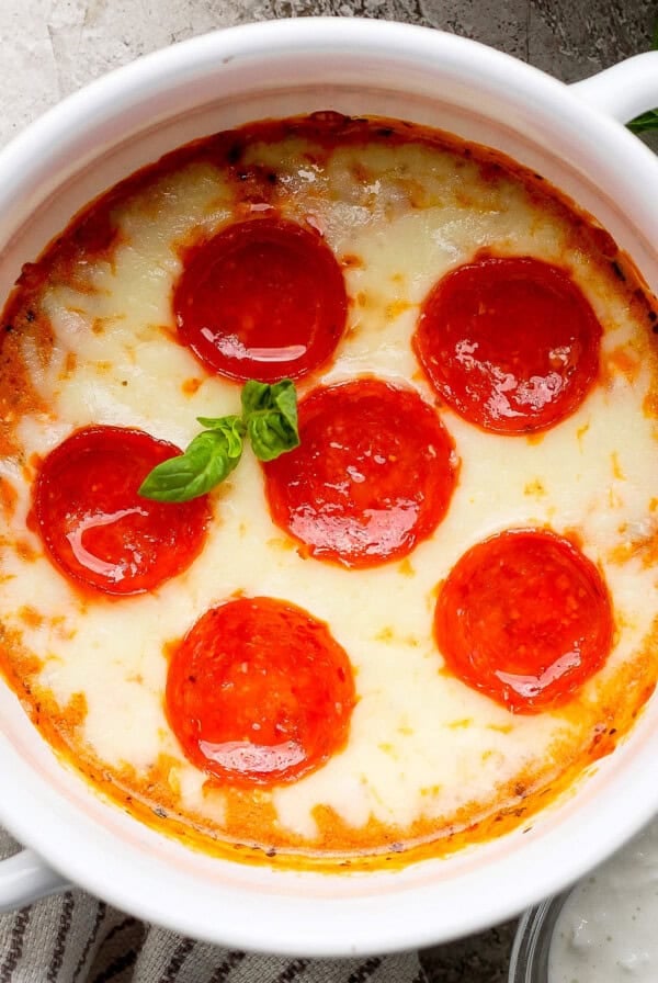 Baked dish with melted cheese and six pepperoni slices, garnished with basil, in a white ceramic bowl on a textured surface.
