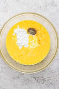 A glass bowl with beaten eggs, baking powder, and black pepper on a light textured surface.
