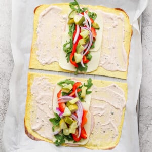 Two open-faced flatbread sandwiches on a baking sheet, with cheese, arugula, red onions, red peppers, and pickles, topped with a creamy spread, placed on parchment paper.