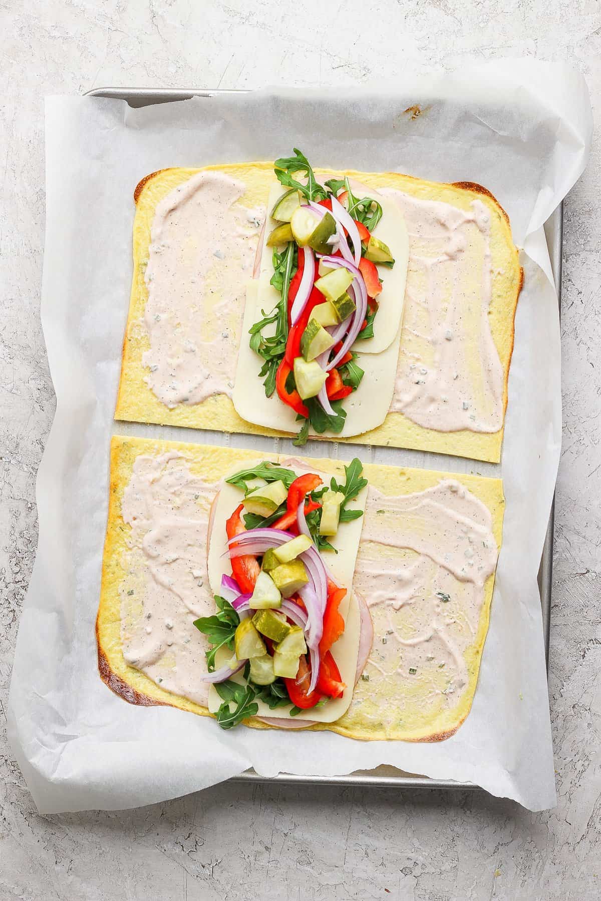 Two open-faced flatbread sandwiches on a baking sheet, with cheese, arugula, red onions, red peppers, and pickles, topped with a creamy spread, placed on parchment paper.