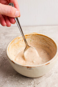 Hand holding a spoon in a ceramic bowl of creamy sauce on a gray surface.