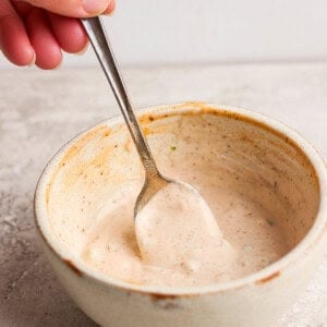 Hand holding a spoon in a ceramic bowl of creamy sauce on a gray surface.