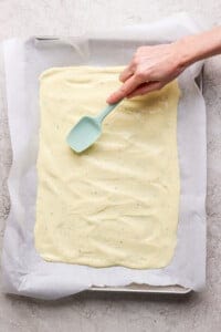 Hand spreading batter evenly on a baking sheet lined with parchment paper using a teal spatula.
