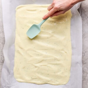 Hand spreading batter evenly on a baking sheet lined with parchment paper using a teal spatula.