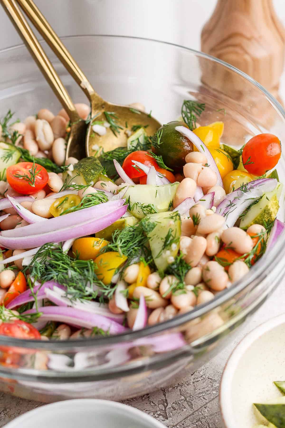 A bowl of vegetable salad with cherry tomatoes, cucumber, white beans, red onion, and fresh dill, tossed with two gold utensils.