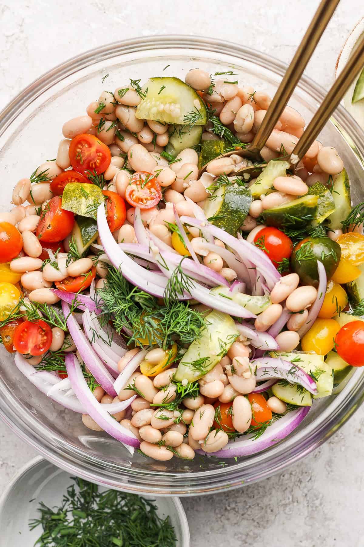 A bowl of bean salad with white beans, cherry tomatoes, red onion, cucumber, fresh dill, and dressing, garnished with herbs, with serving utensils.