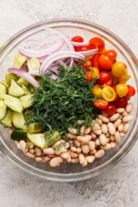 Glass bowl with sliced red onions, halved cherry tomatoes, cucumber chunks, white beans, and a mound of fresh dill.