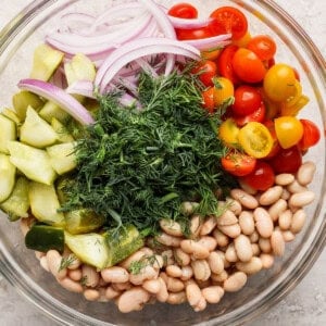 Glass bowl with sliced red onions, halved cherry tomatoes, cucumber chunks, white beans, and a mound of fresh dill.