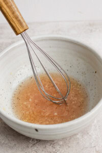 A metal whisk rests in a white bowl filled with a light brown liquid, possibly a vinaigrette or sauce, set on a textured surface.