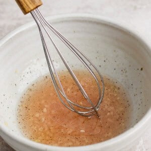 A metal whisk rests in a white bowl filled with a light brown liquid, possibly a vinaigrette or sauce, set on a textured surface.